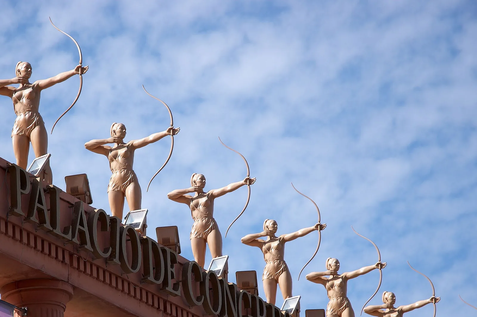 Statues of archers drawing bows along the roof of the Palacio de Congresos, above its lettering, against a blue sky with thin cloud