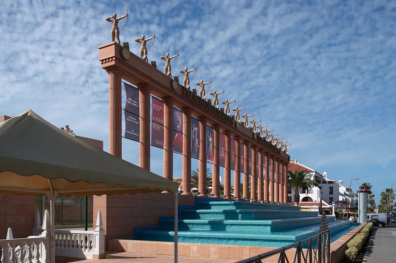 The colonnade of the Palacio de Congresos in daylight, with a row of archer statues on the roof, tall columns hung with banners and stepped turquoise pools at its foot