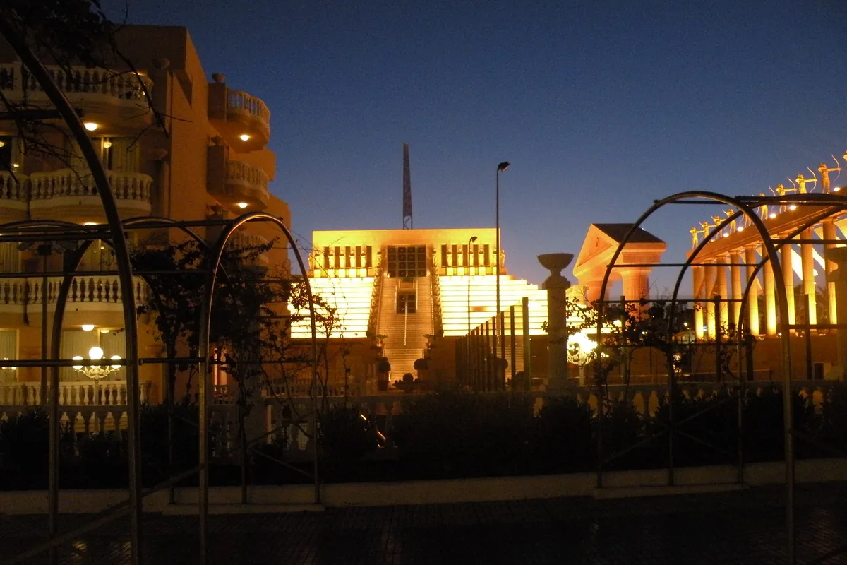 A stepped building lit gold at night beside the Palacio de Congresos colonnade, with hotel balconies on the left and a dark sky above