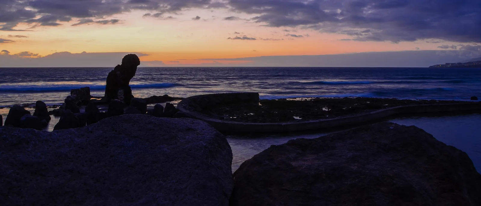 The sun setting over the Atlantic at Playa de las Américas, with an orange and purple sky, a line of surf, and dark rocks and a stone sea wall in the foreground